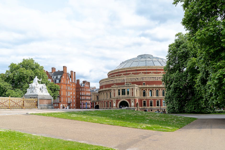 An outdoor scene near Alexandra Palace features a green lawn with a paved pathway leading towards the historic building's entrance. To the left, there is a stone sculpture depicting figures, situated on a pedestal, adjacent to a decorative metal fence with patterned panels. Behind the sculpture, mature trees with lush foliage partially conceal a row of red-brick residential buildings. The main Alexandra Palace building, with its distinct round shape, red brick facade, and arched windows, dominates the background, topped by a large, domed roof with a glass or metal covering. The sky above is partly cloudy with patches of blue visible. The image captures the peaceful outdoor environment that may be encountered during a house or office move in the Alexandra Palace area, reflecting the importance of proximity to landmarks during relocation planning. The presence of the outdoor environment and surrounding architecture aligns with the services provided by Man with Van Fortis Green for furniture transport, packing, and home relocation processes.
