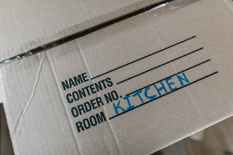 Close-up of a cardboard moving box with handwritten blue text indicating it contains kitchen items and is designated for the kitchen room, with printed labels showing sections for name, contents, and 