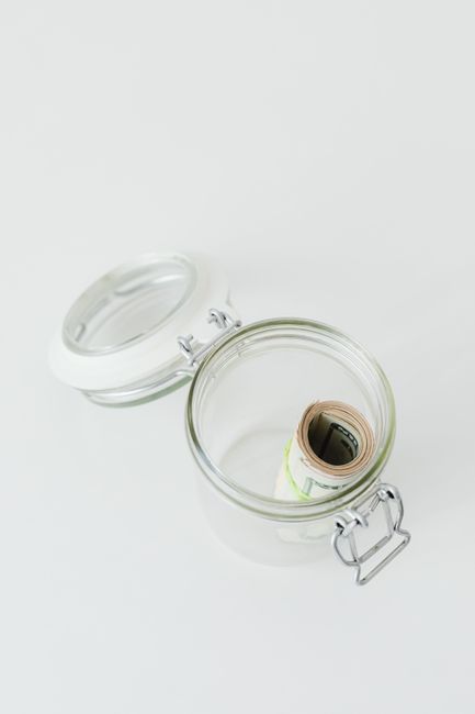 A clear, empty glass jar with a metal clasp and a rubber sealing gasket sitting on a white surface. The jar's lid is open and positioned slightly behind it. Inside the jar, there is a rolled-up document or banknote. The setting is minimalistic with no other objects visible, emphasizing the jar's transparency and the details of the sealing mechanism, representing a clean and organized approach to packing materials often involved in home relocation and removals by Man with Van Fortis Green, serving the Fortis Green area for end-of-lease clearance and deposit-saving tasks.