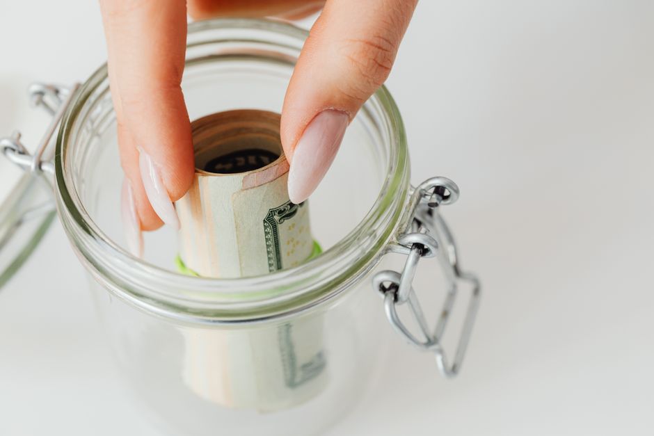 A close-up view of a person's hand with manicured nails placing a rolled banknote into a transparent glass jar with a metal latch and sealing mechanism, possibly for savings or deposits. The jar is situated on a plain white surface, and part of the metal latch and rubber seal are visible beside it. The image conveys themes of financial management or savings, relevant to house removal services like end-of-lease clearance. In the context of a home relocation or furniture transport, this visual could symbolize deposit savings or budget planning associated with moving or clearance procedures conducted by companies such as Man with Van Fortis Green, as mentioned on the webpage titled 'End-of-lease clearance in Fortis Green.'