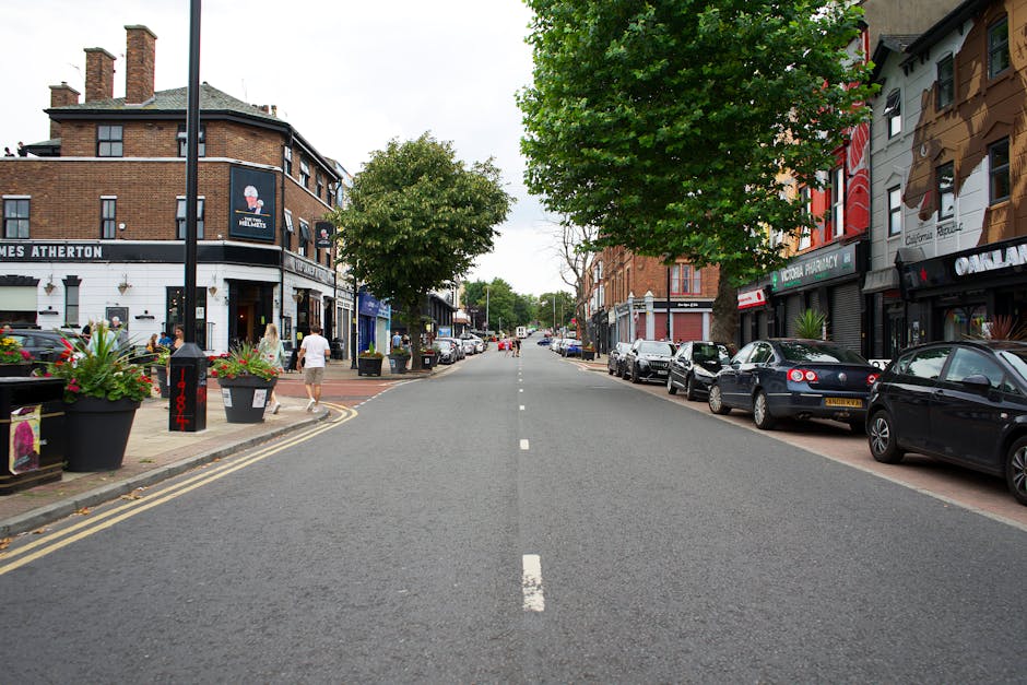 A straight view of a residential street in Fortis Green featuring parked cars along both sides of the road and a mix of commercial and residential buildings with brick and painted facades. The street is flanked by trees with lush green foliage, providing partial shade to the pavement and parked vehicles. In the foreground on the pavement, there are large black planters filled with colorful flowers and a black bollard near the curb, indicating a designated parking or loading area. People are walking along the sidewalk, some carrying shopping bags or talking. The scene is during daytime with natural light, and the street appears quiet, typical of a local shopping and residential area. The image is relevant to [PAGE_TITLE], illustrating urban environment conditions for house removals or furniture transport, with visible arrangements for parking and loading that could assist logistics planning for a move as coordinated by [COMPANY_NAME], focusing on efficient and accessible home relocation services.