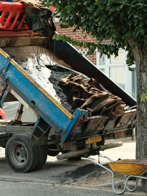 A small blue flatbed truck parked on a residential street, with its bed tilted to unload dark asphalt into a yellow wheelbarrow. The asphalt is partially covered by a brown cloth, and the truck's license plate reads L119 AFE. In the background, there is a red brick house with white-framed windows and a large leafy tree providing partial shade. The scene depicts construction or repair work likely related to home relocation or building maintenance, with the truck positioned near a pavement area adjacent to the house. This image illustrates the typical process involved in site preparation or groundwork associated with house removals or renovations, supported by the presence of equipment such as the wheelbarrow and the asphalt mixture. Man with Van Fortis Green offers professional removals services, including site clearing and transportation, as implied by the context.