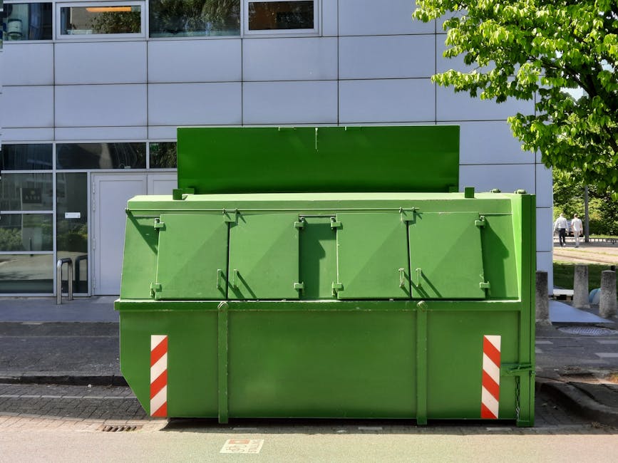 A large green waste or recycling skip positioned on the pavement outside a modern commercial building with a grey facade and large windows. The skip is made of metal and features multiple hinged lids on the top, secured with latch mechanisms. It has red-and-white reflective strips on its lower corners for visibility. The skip is empty, with no visible contents, and is set on a flat paved surface next to a street, with a tree and some pedestrians visible in the background. The scene is illuminated by daylight, indicating normal outdoor working conditions. This image reflects the type of equipment used during home relocation and furniture transport, particularly for disposing of waste or debris during moving or packing processes, as part of professional removals by companies such as Man with Van Fortis Green.