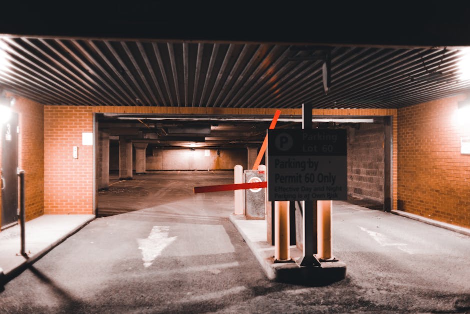 An underground parking garage with brick and concrete walls, featuring a gated entrance with red and white barrier arms in a lowered position. A parking sign indicating 'Parking Lot 60' and 'Permit 60 Only' is mounted on a black post near the barrier. The floor is paved with asphalt and has a white directional arrow painted on it, indicating the flow of vehicle traffic. Inside the garage, various concrete pillars support the ceiling, which is covered with dark, slatted panels. The lighting is dim, with a few bright wall-mounted fixtures providing illumination. This setting suggests a controlled parking area where professional movers from Man with Van Fortis Green may load or unload furniture and boxes during a home relocation process, highlighting the logistics of furniture transport and packing during house removals.