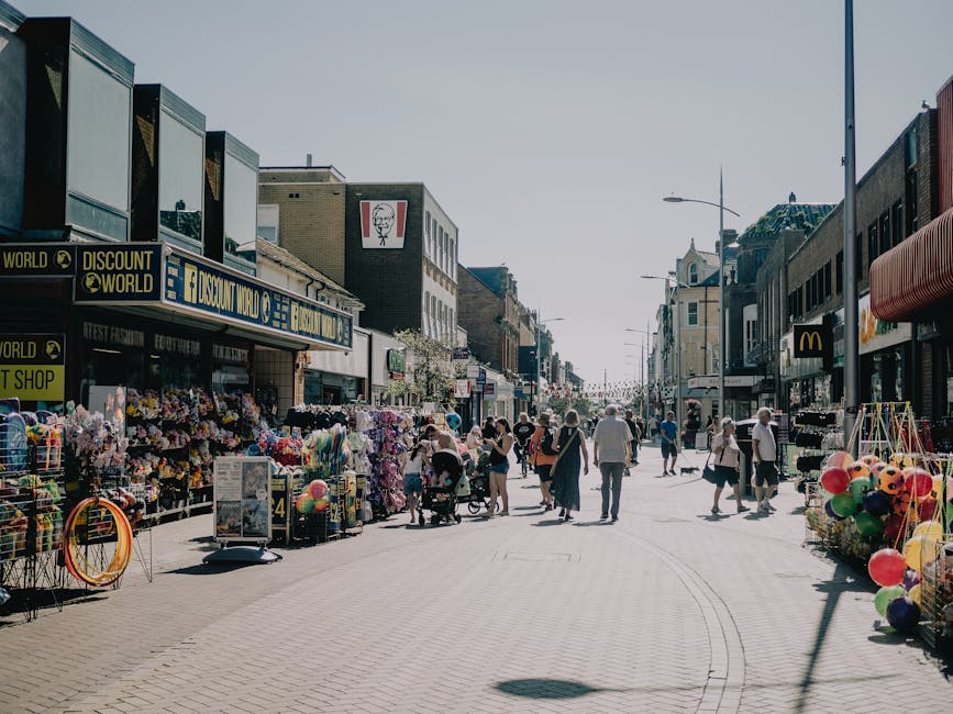 A busy street scene on Muswell Hill Broadway during daylight hours, with pedestrians walking along the pavement and several individuals visible carrying boxes and small furniture items as part of a home relocation or moving process. On the left side, outdoor market stalls display an array of colorful plastic toys, balloons, and packaged goods, with some items stacked on carts and tables. The storefronts include signs for businesses such as 'Discount World' and a fast-food outlet, with signage and window displays visible. On the right side, more shops and storefronts line the street, which is illuminated by streetlights and natural sunlight, contributing to the bustling atmosphere. The pavement is made of light-colored bricks, and there are a few street fixtures, like waste bins and signage, scattered throughout. The scene captures the movement and activity typical of a vibrant shopping district, conveying the context of a busy commercial area that could also serve as a location for coordinating moving or furniture transport activities, as indicated by the presence of people handling packing and loading items.