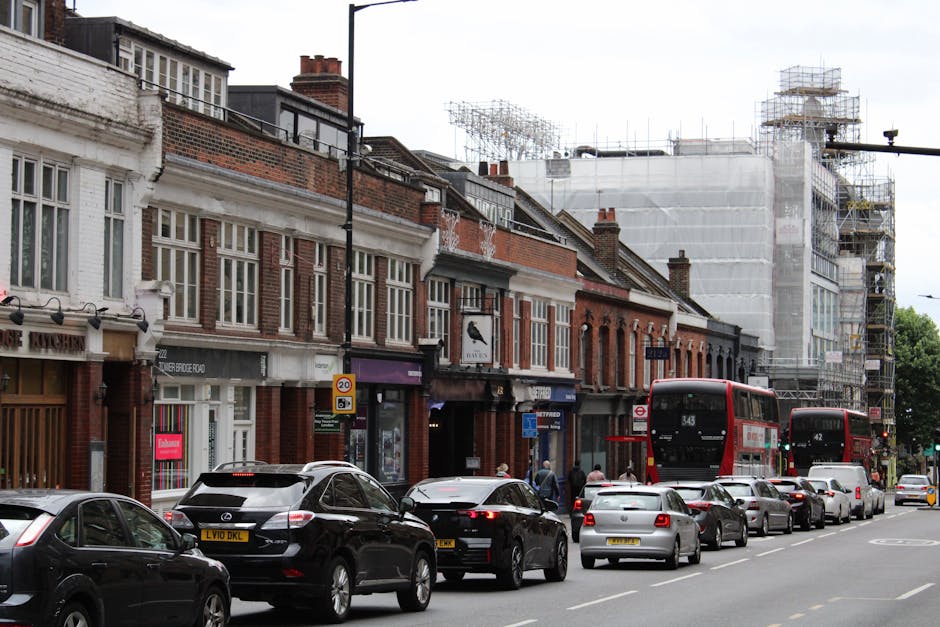 A busy street scene in Muswell Hill Broadway during a home relocation, featuring a row of brick and white-painted terraced buildings with retail shops on the ground floors and residential units above. Several parked cars, including a black Mercedes and a silver hatchback, line the roadside pavement. In the background, scaffolding covers a building under construction or renovation, indicating ongoing building work. Two red double-decker buses are stopped or moving along the street, while pedestrians walk on the sidewalk, some near the entrance of shops. The image captures the typical urban environment for house removals and furniture transport, with visible materials such as cardboard boxes, plastic wrapping, and wrapped furniture likely being prepared for loading. Man with Van Fortis Green may use such scenes to illustrate the logistical flow involved in packing, loading, and transportation during a house removal within London’s bustling streets.