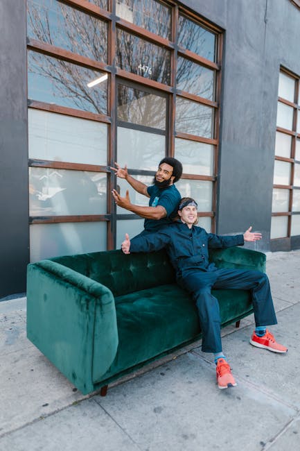 Two men are outside a building with large, grid-like wooden window frames and frosted glass panes, preparing for a home relocation. One man, dressed in dark clothing, is seated on a green velvet sofa positioned on the sidewalk, with his arms extended outward in a welcoming gesture. The other man, wearing a blue t-shirt and jeans, stands behind the sofa, smiling and giving a thumbs-up while holding onto the window frame with one hand. The scene suggests a casual moment during packing or furniture transport, with natural daylight illuminating the area, which is typical during a moving process. The presence of the sofa on the pavement indicates it is in the process of being loaded into or unloaded from a van for a house move, consistent with the services of Man with Van Fortis Green, specialising in removals and furniture transport in the area.