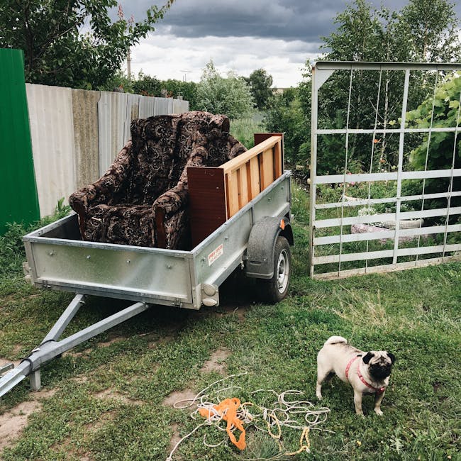 A small outdoor area with a metal flatbed trailer attached to a vehicle, positioned on a grassy surface next to a fenced garden with a wooden trellis and various plants. The trailer is loaded with a large, irregularly shaped piece of wood or bark, along with a small wooden panel and a grey plastic container. Nearby, a pug dog with a pink collar stands on the grass, surrounded by orange and beige ropes or straps, indicating a home relocation or furniture transport process. Overhead, dark storm clouds create a moody sky, and in the background, there are trees and bushes, suggesting a residential setting. This scene captures the phasing of packing and moving furniture or bulky items during a move, possibly organized by Man with Van Fortis Green for house removals or similar services.