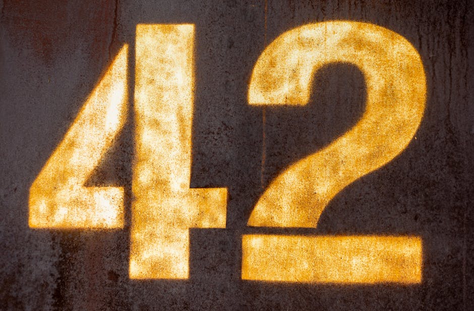 The image shows a close-up of a textured, dark surface illuminated by a warm, yellowish light projecting the digits '42' in large, bold font. The lighting creates a glowing effect around the numbers, highlighting their edges and the rough surface details. The number '4' is positioned on the left, with its vertical and diagonal lines clearly visible, while the number '2' appears to the right, with its curved and straight components. The scene suggests an outdoor or indoor setting with minimal background visible, focusing attention on the illuminated numerals, which are likely part of a house number or an area marker. This type of lighting and presentation can relate to home or building identification in a professional moving or relocation context, as provided by companies like Man with Van Fortis Green, involved in packing, furniture transport, and home relocation services.