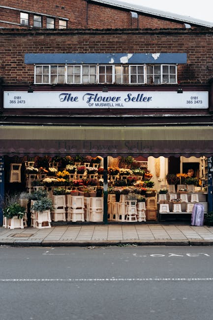 The image shows the storefront of 'The Flower Seller of Muswell Hill', a flower shop situated beneath a brick building. The shop has a maroon awning with the business name and contact details, and a l